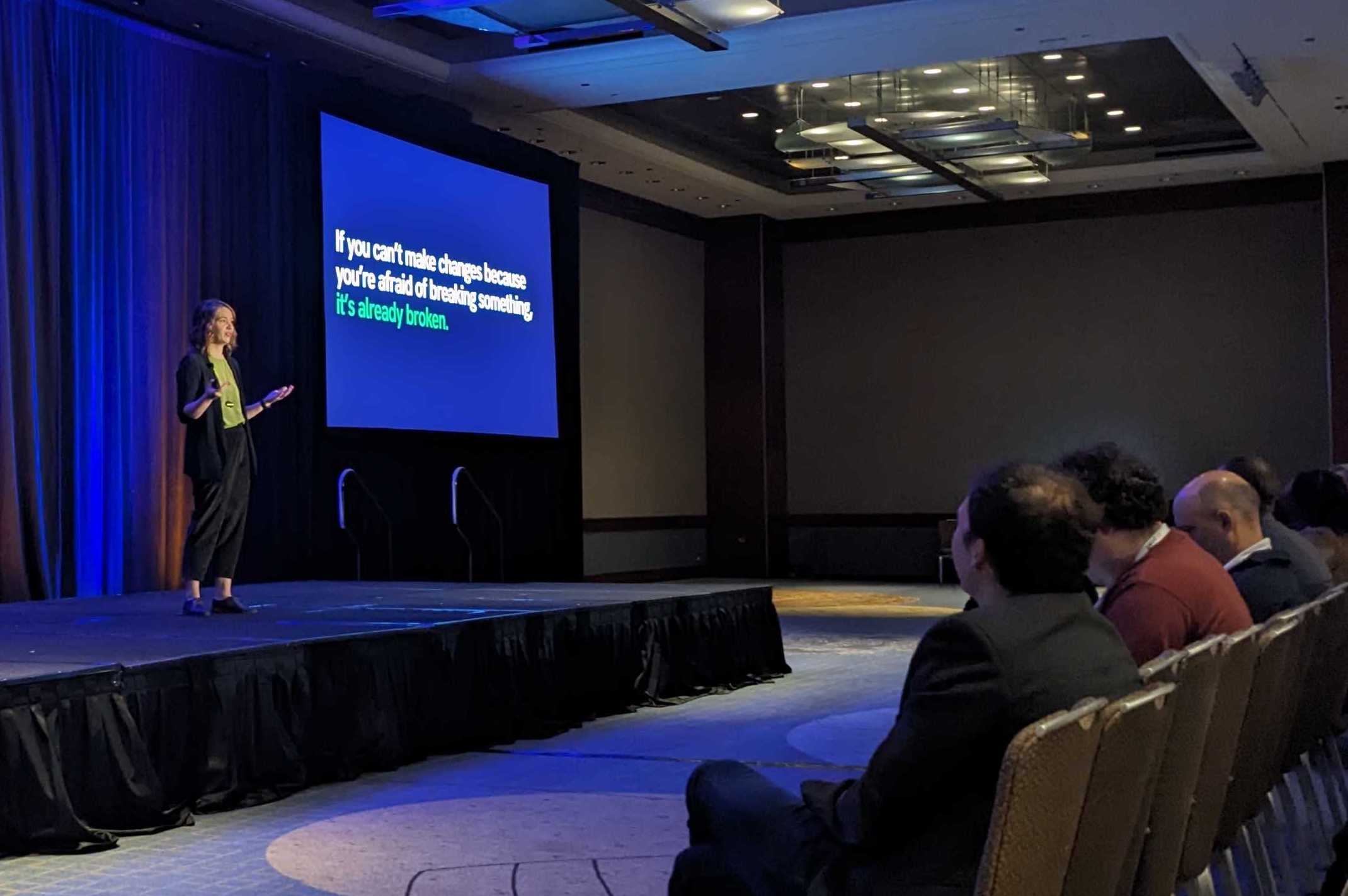 photo of Kara Woo on stage beside screen projecting text "if you can't make changes because you're afraid of breaking something, it's already broken". Several seated audience members visible in front row.